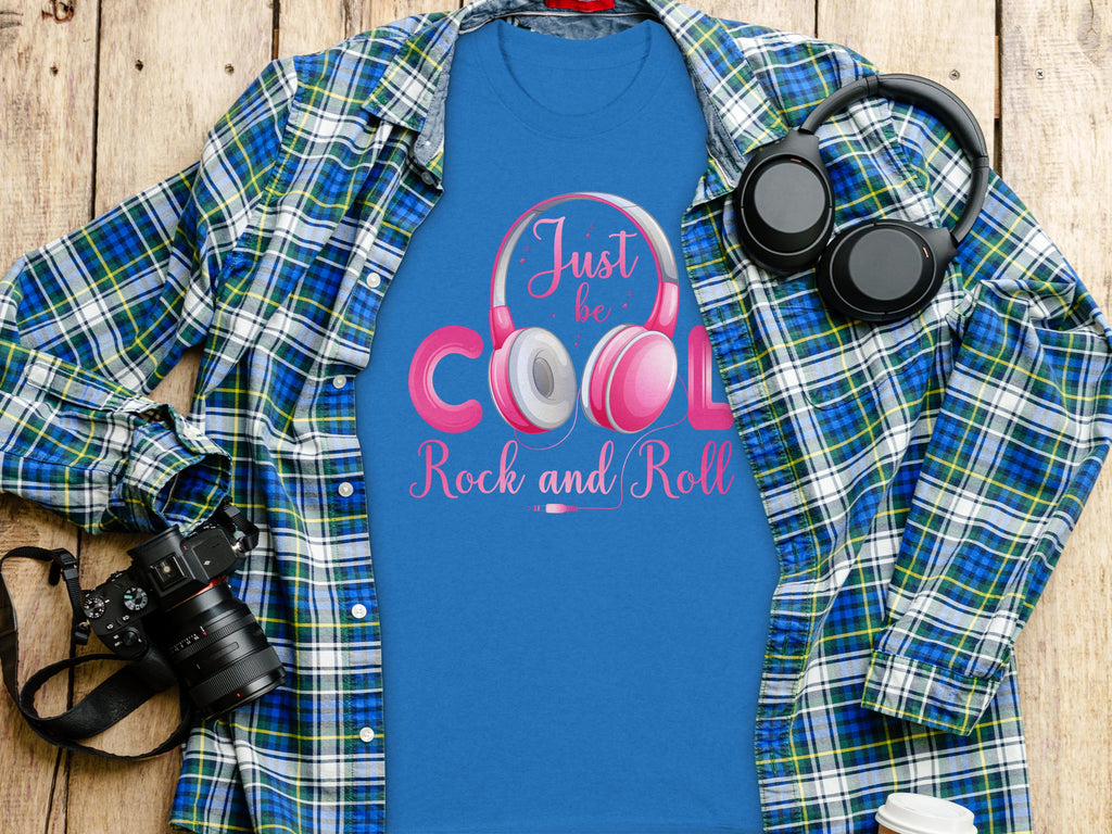 Garment Graphics blue t-shirt features Just be cool, rock and roll in pink and white with playful headphones. Its styled on a plaid shirt next to black music lovers headphones, a camera, and a disposable coffee cup on a wooden surface.
