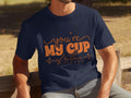 Sitting on a wooden bench outdoors, a person wears Garment Graphics 100% cotton T-shirt with playful orange text Youre My Cup of Tea and decorative elements. Soft focus grass and trees create a serene backdrop.