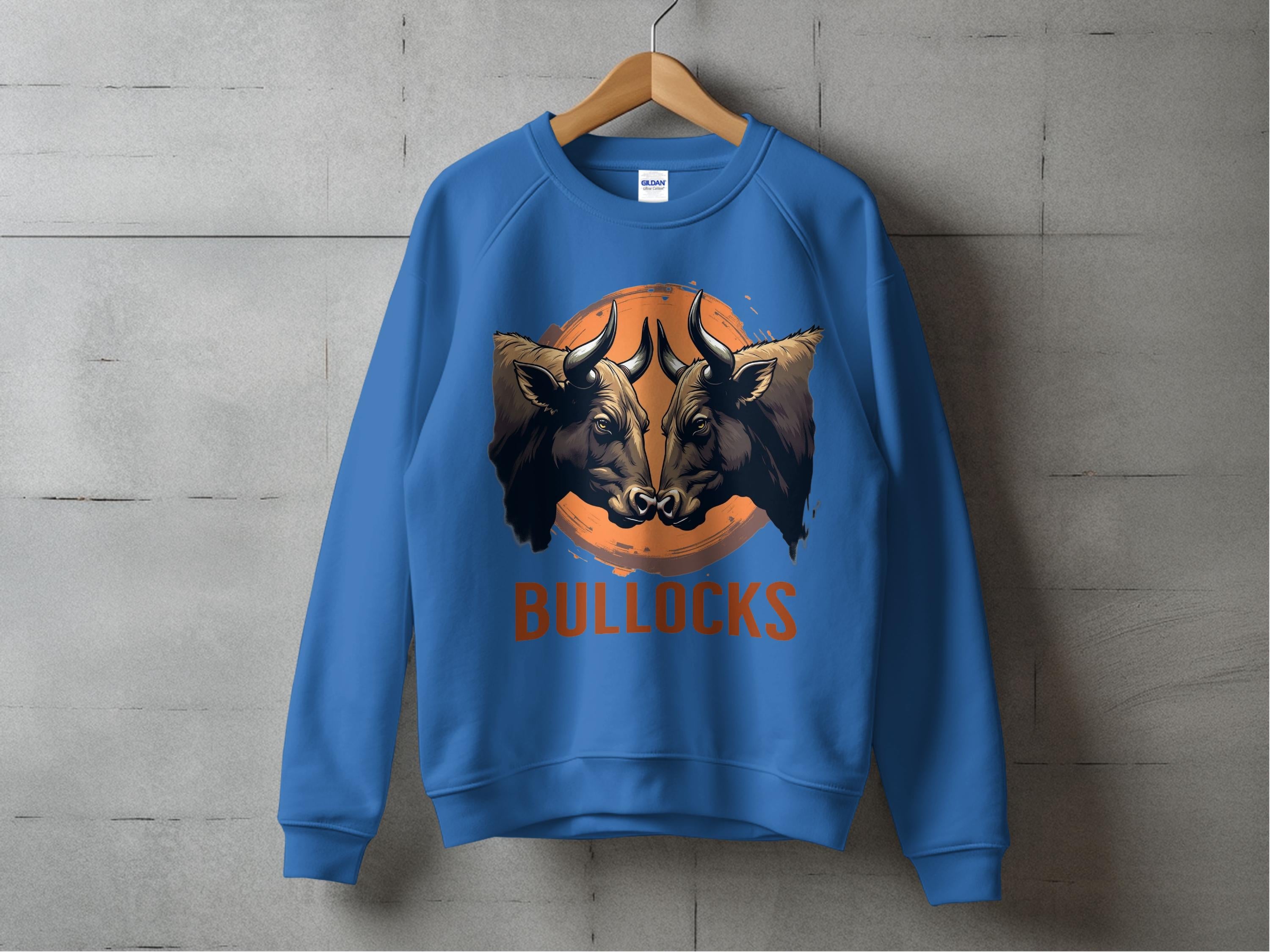 A loose-fit blue sweatshirt featuring a bullocks graphic of a bulls head in an orange circle hangs on a wooden hanger against a concrete wall. The bold BULLOCKS text enhances its allure. Brand: Garment Graphics.