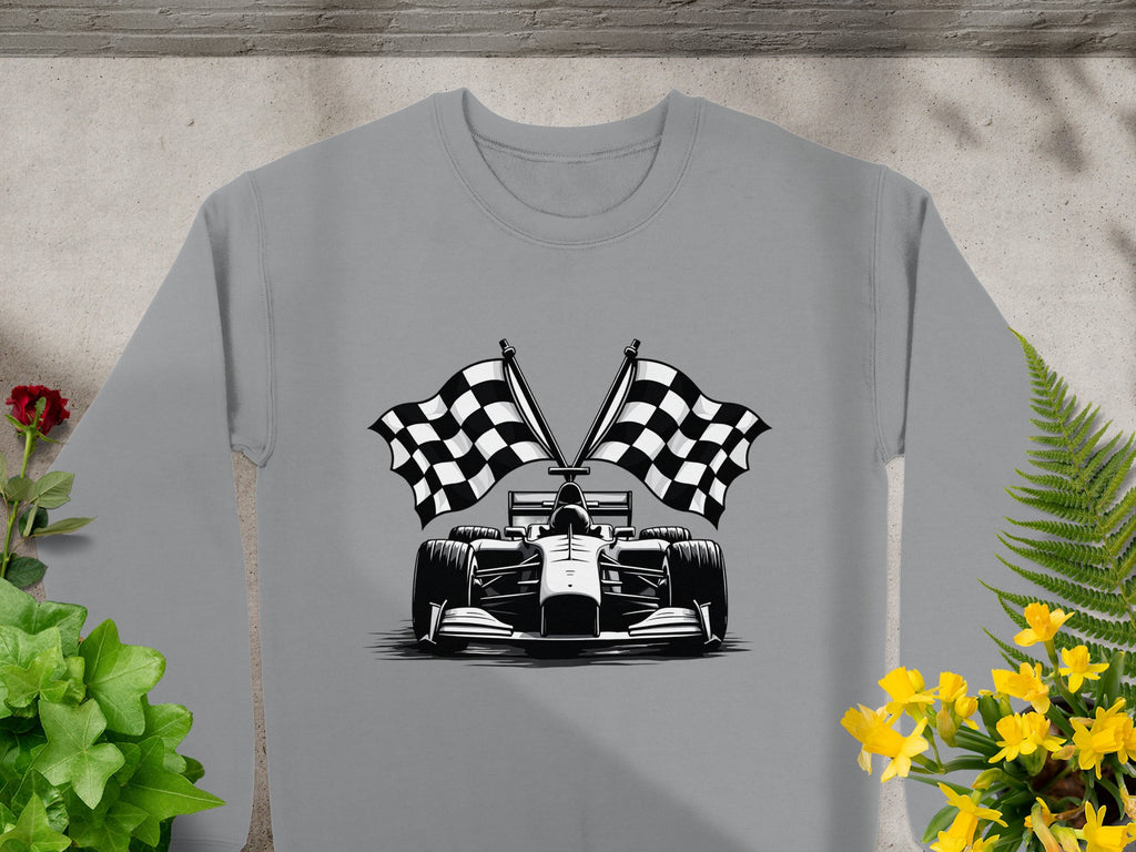 The Garment Graphics gray sweatshirt features a striking Formula 1 car and crossed checkered flags graphic, laid on concrete amidst green plants and vivid yellow and red flowers.
