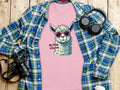 A pink Llama T-Shirt by Garment Graphics, featuring a llama with sunglasses and the text No Prob-Llama, lies on wood surrounded by a blue-green flannel, camera, headphones, and glasses. Ideal for family sizes.