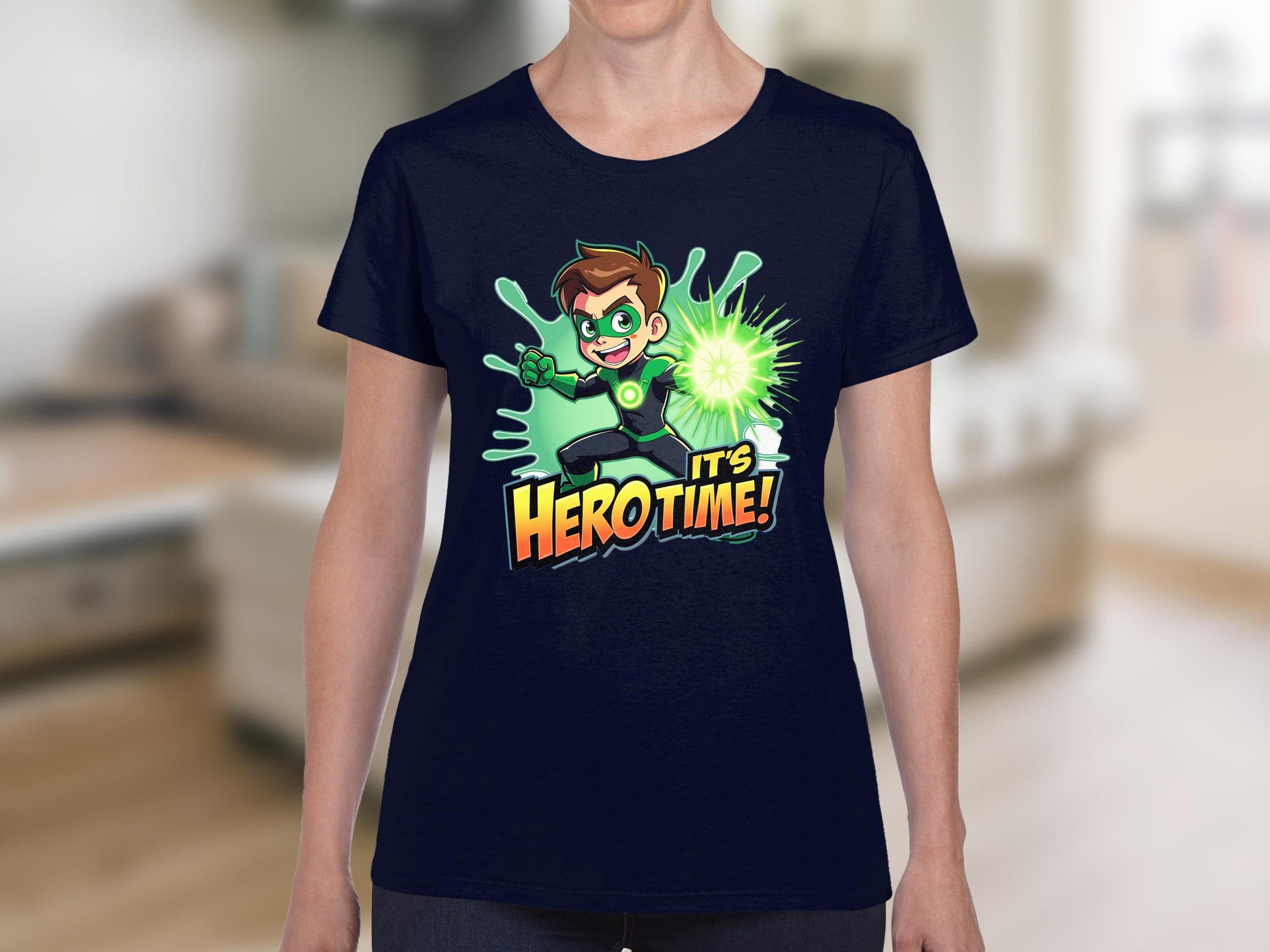 A person wearing a Garment Graphics navy blue T-shirt displays a cartoon character with spiky hair holding a glowing green sphere, embodying a green superhero. The text Its Hero Time! enhances the standout effect against the blurred indoor background.