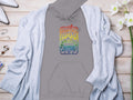 Garment Graphics retro gray hoodie boasts a vibrant soda can graphic with The Doors 1969. On a wooden surface, it stylishly pairs with a light gray cardigan, white heels, jewelry, and white flowers. Ideal for those embracing Woodstock 1969 vibes.