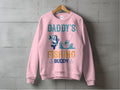 A pink graphic sweatshirt by Garment Graphics hangs on a wooden hanger against a concrete wall, ideal for outdoor enthusiasts. It showcases the playful Daddys Fishing Buddy design with a cartoon fish, someone fishing from a boat, and decorative elements.