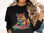 A person with long wavy blonde hair indoors is wearing a Garment Graphics black sweatshirt featuring a colorful big cat design. The cougar graphic in blue, orange, yellow, and pink emphasizes its unique style. The unisex sizing offers versatile wear.