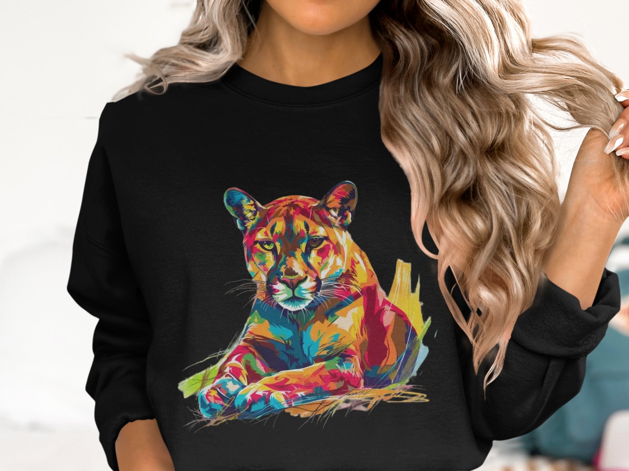 A person with long wavy blonde hair indoors is wearing a Garment Graphics black sweatshirt featuring a colorful big cat design. The cougar graphic in blue, orange, yellow, and pink emphasizes its unique style. The unisex sizing offers versatile wear.