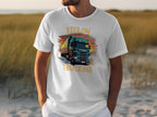 Someone wearing a white Garment Graphics shirt featuring a big green truck and the phrase KEEP ON TRUCKING stands outside among sandy ground and tall grasses, an ideal backdrop for truck lovers.