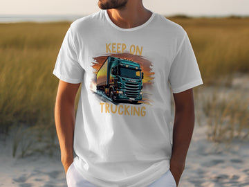 Someone wearing a white Garment Graphics shirt featuring a big green truck and the phrase KEEP ON TRUCKING stands outside among sandy ground and tall grasses, an ideal backdrop for truck lovers.