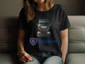 Seated on a couch, a person wears a black Garment Graphics T-shirt with a Scania truck illustration and blue logo. Their head is unseen, with one hand resting on their knee, highlighting their love for all things automotive.