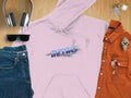 Flat lay featuring Garment Graphics clothing and accessories: trendy headphones, sunglasses, blue jeans, a pink Retro hoodie with vivid colors, an orange button-up shirt with additional sunglasses, a wristwatch, and a set of keys on a wooden surface.