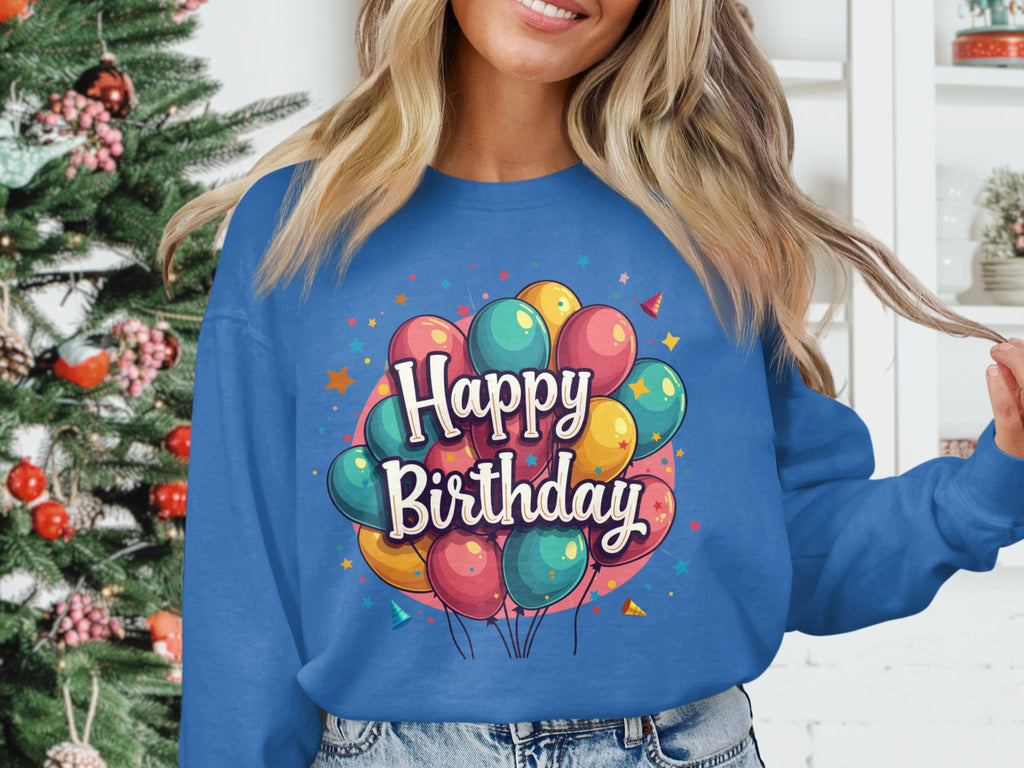A person stands by a Christmas tree in a vibrant blue Garment Graphics sweatshirt, featuring colorful balloons and Happy Birthday text. This festive unisex piece is perfect for any occasion.