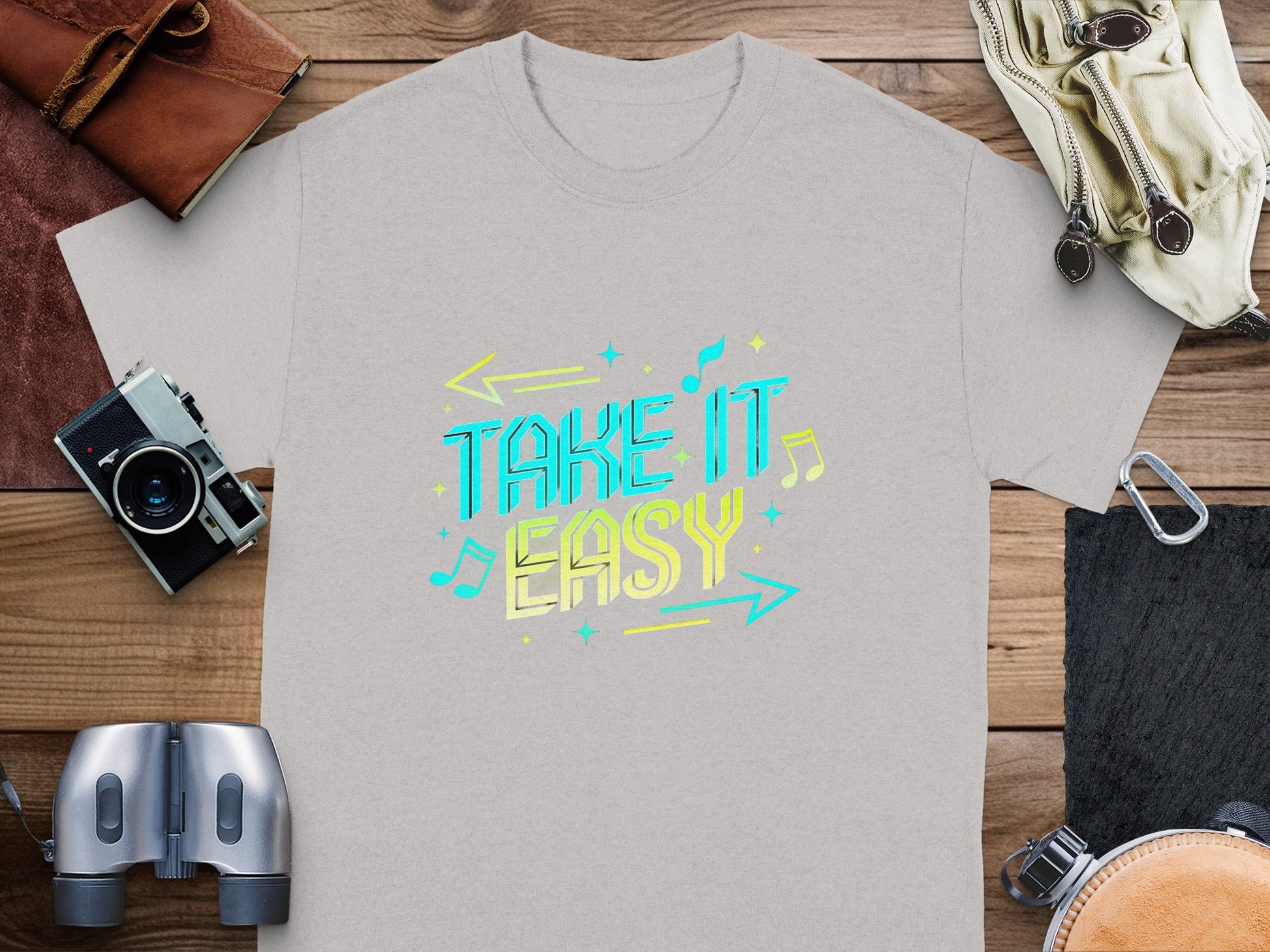 The Garment Graphics gray T-shirt features vibrant Take It Easy text in colorful letters with arrows, musical notes, and stars. Its displayed on a wooden surface alongside a camera, leather pouch, binoculars, and gloves.