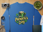 A vibrant blue sweatshirt from Garment Graphics features a festive Happy Patricks Day design with a green mug filled with foam and shamrocks, alongside a camera, lens, sliced kiwi in a bowl, map, dark green book, and burlap sack on the wooden surface.