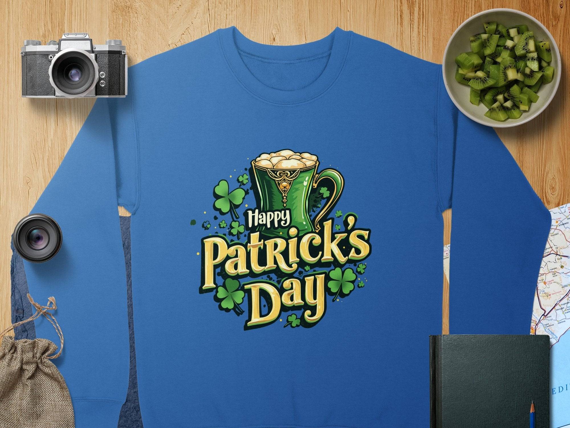 A vibrant blue sweatshirt from Garment Graphics features a festive Happy Patricks Day design with a green mug filled with foam and shamrocks, alongside a camera, lens, sliced kiwi in a bowl, map, dark green book, and burlap sack on the wooden surface.