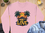 Garment Graphics One Love Sweatshirts in pink showcases a sunset with palm trees, an airplane, and mountains. This unisex sweatshirt is styled on wood beside a latte and jacket, ideal for relaxed days.