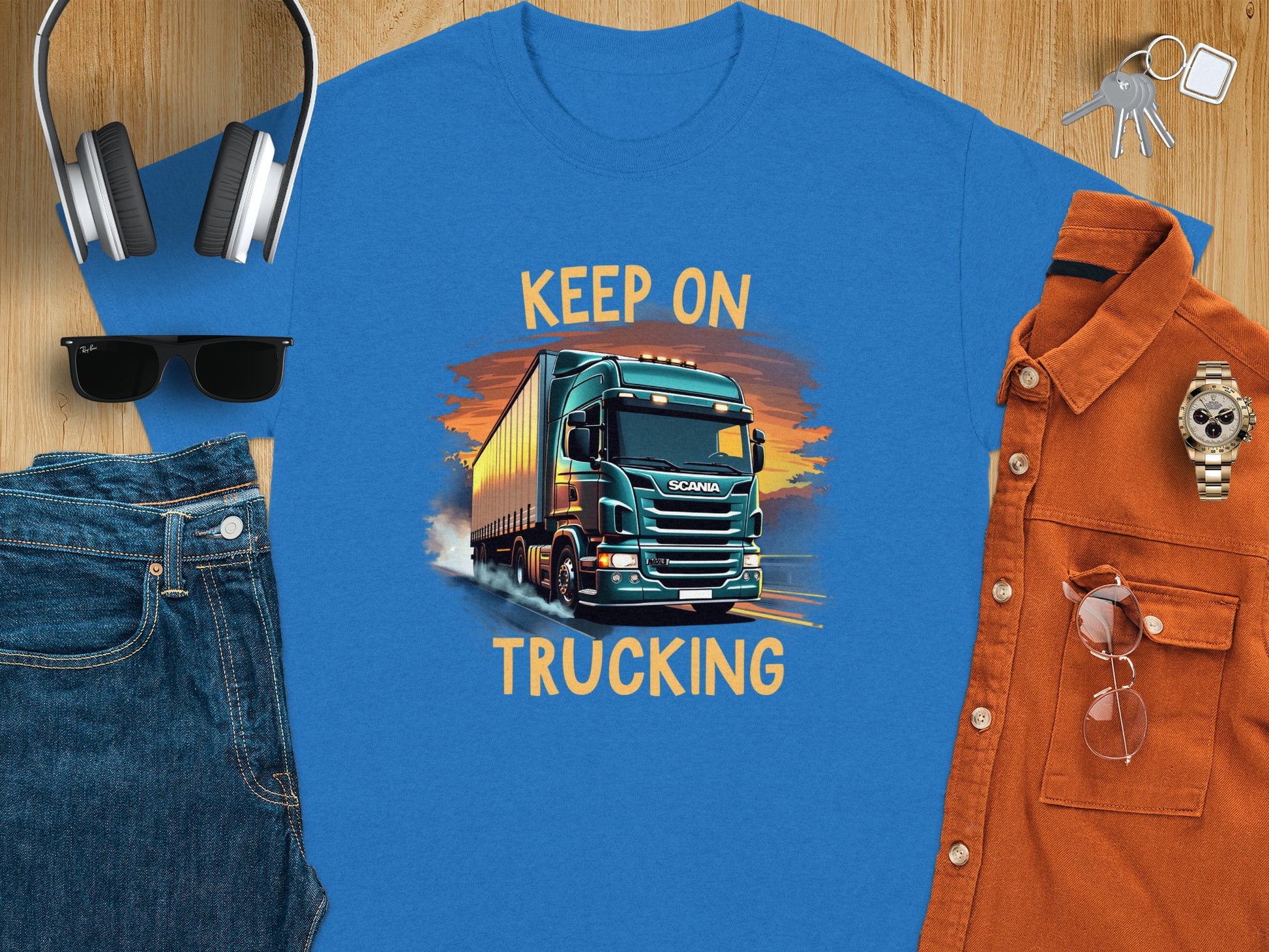 The Garment Graphics blue graphic t-shirt, showcasing a truck with Keep on Trucking, is ideal for truck fans. Pair it with essentials like headphones, a smartphone, keys, sunglasses, jeans, an orange shirt with glasses, and a watch on rustic wood for a cool vibe.
