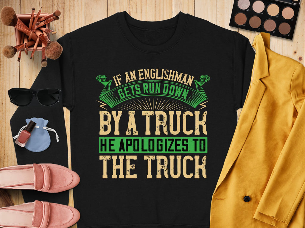 The Garment Graphics black sweatshirt showcases a bold, humorous green and beige text: If an Englishman gets run down by a truck, he apologizes to the truck. Styled unisex on wood with accessories like makeup, sunglasses, and clothes.