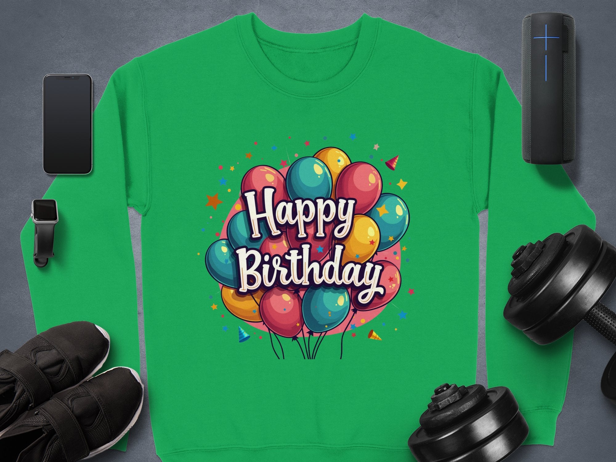 The Garment Graphics green sweatshirt, featuring a colorful balloon design and Happy Birthday text, is ideal for celebrations. Unisex in size, it pairs well with smartphones, smartwatches, speakers, black sneakers, and dumbbells on a dark surface.