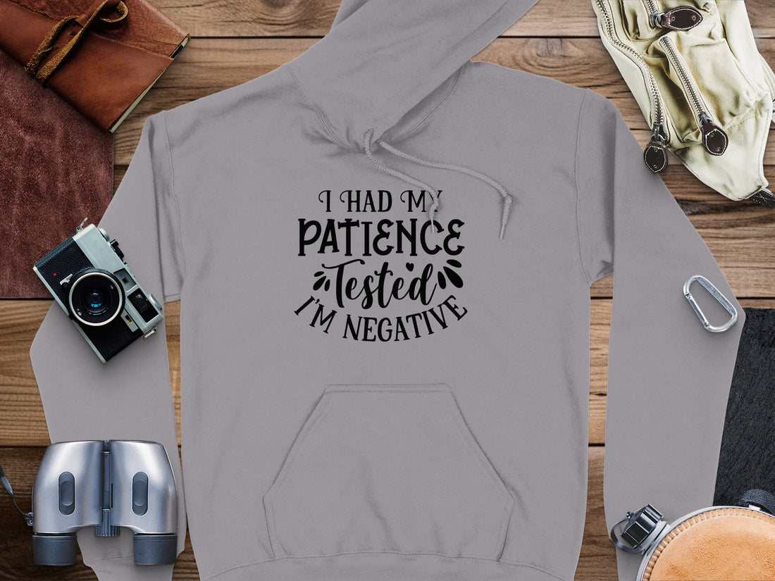 The gray I had my patience tested, Im negative novelty hoodie by Garment Graphics features a funny statement design. Its shown with a camera, binoculars, leather pouch, strap, and backpack on a wooden surface. Perfect for those who enjoy humorous hoodies.