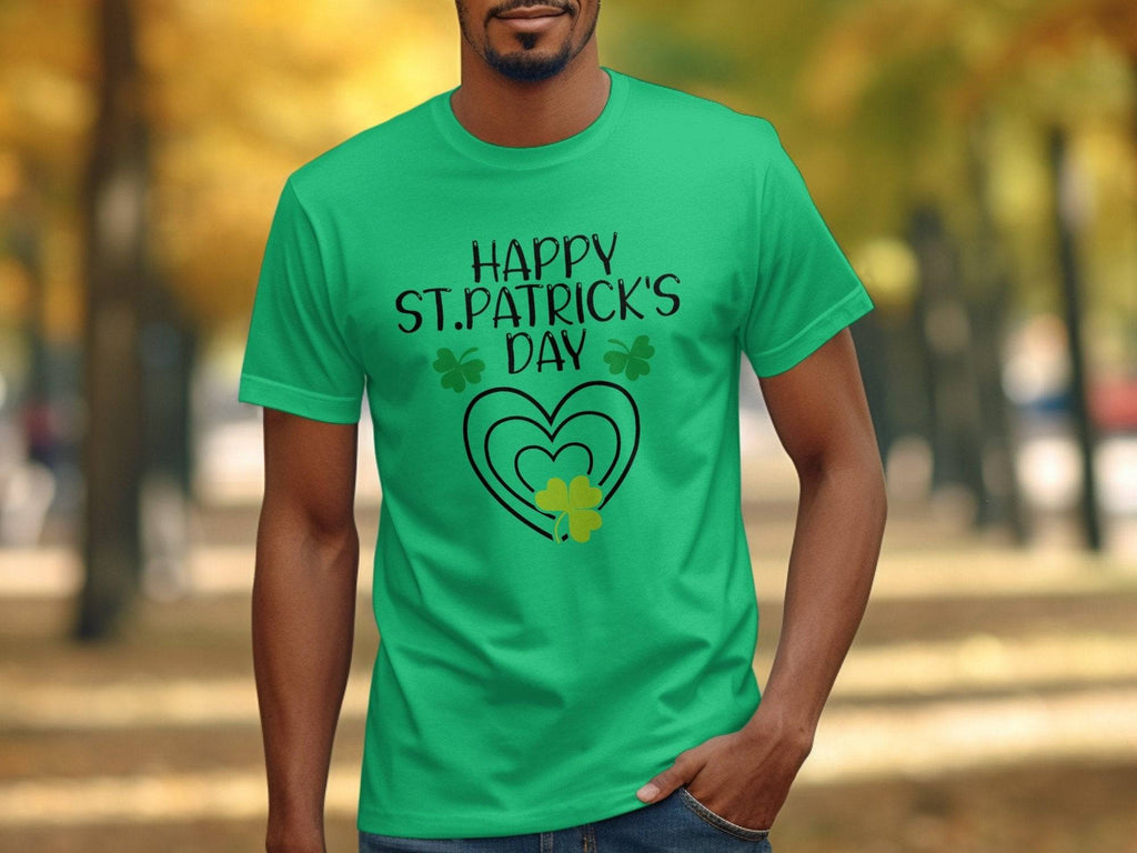 A person wearing a Garment Graphics green T-shirt featuring Happy St. Patricks Day with hearts and shamrocks is outdoors, framed by blurry autumn trees in the background.