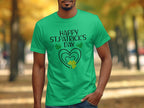A person wearing a Garment Graphics green T-shirt featuring Happy St. Patricks Day with hearts and shamrocks is outdoors, framed by blurry autumn trees in the background.