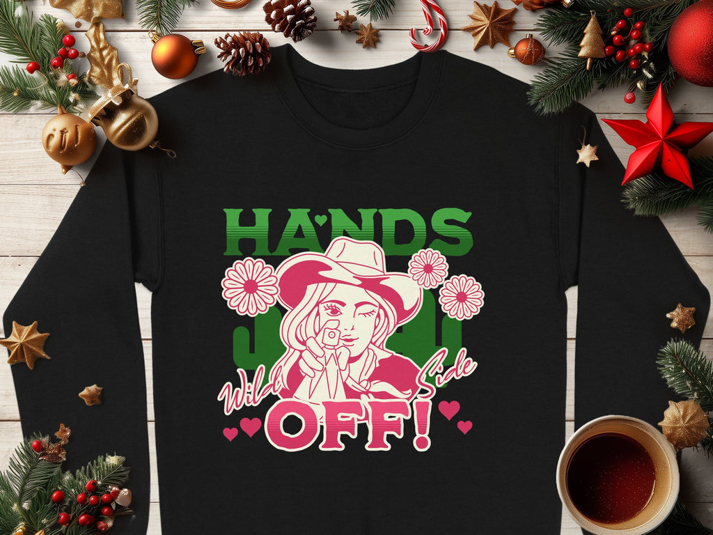 Garment Graphics black loose-fit sweater features a cowboy hat graphic with flowers and hearts, alongside HANDS OFF Wild Sale in green, pink, and white. It is styled against a wooden background with Christmas decorations and a coffee cup to highlight its unique charm.