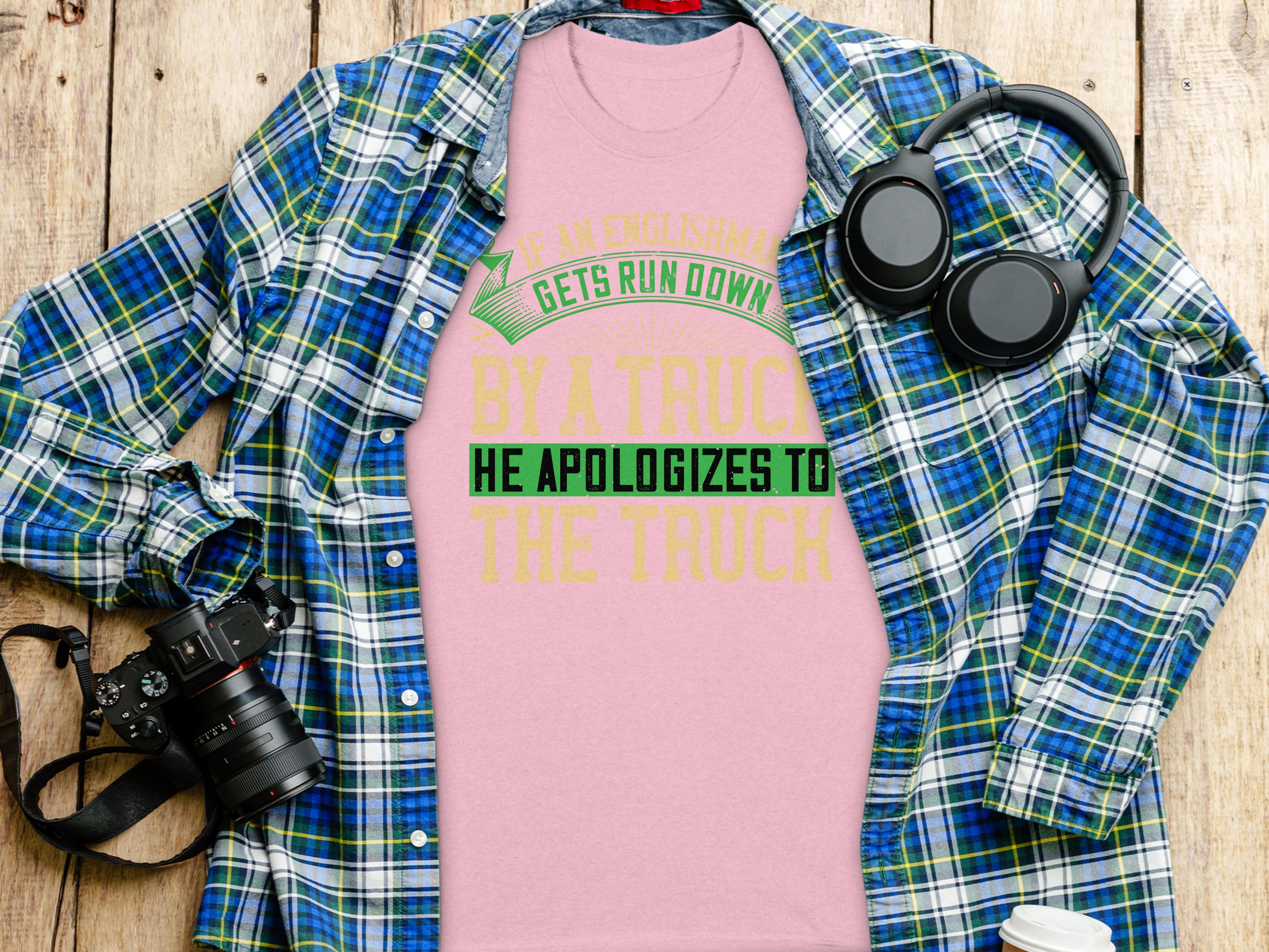 A Garment Graphics pink T-shirt with the witty If an Englishman gets run down by a truck, he apologizes to the truck design is displayed on a wooden surface, surrounded by a blue plaid shirt, headphones, a camera, and a white paper cup.