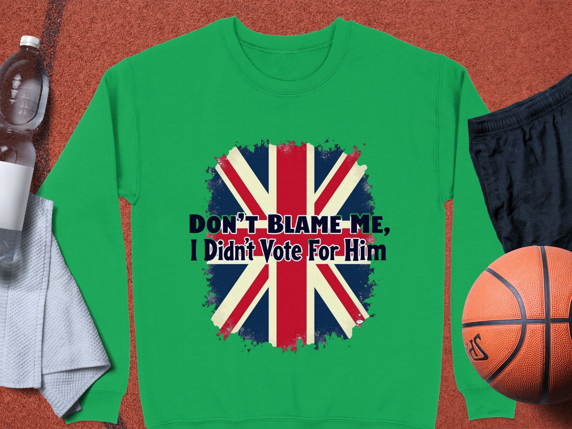 A Garment Graphics green sweatshirt with the British flag and Dont Blame Me, I Didnt Vote For Him design lies on a surface, surrounded by a basketball, black shorts, a water bottle, and a white towel.
