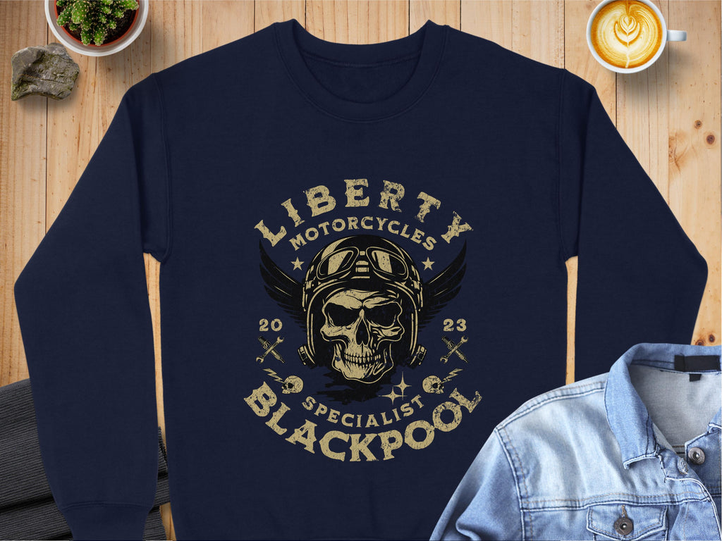 A dark blue sweatshirt from Garment Graphics showcases the biker style with a vintage motorcycle skull graphic, complete with goggles and a winged helmet. The text says Liberty Motorcycles Blackpool Specialist 2023, stylishly placed on a wooden surface beside a denim jacket.