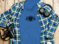 A blue Garment Graphics hoodie featuring a bold Scanir Power logo lies next to a coordinating blue and green plaid shirt, headphones, a camera, and a white cup on wood. This classic fit ensemble exudes style and comfort.