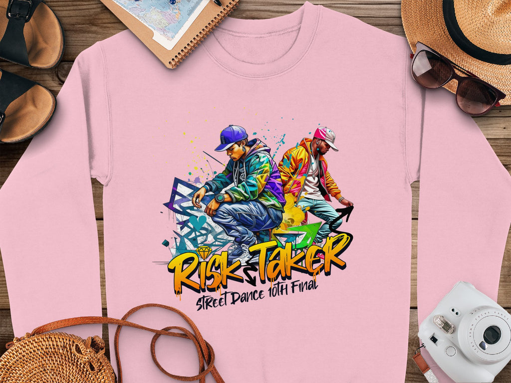 The Garment Graphics pink sweatshirt features a vibrant graphic of two urban dancers celebrating the Risk Taker Street Dance 10th Final with abstract designs, accompanied by stylish sunglasses, a hat, a notebook, and a vintage camera on the wooden surface.