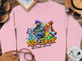 The Garment Graphics pink sweatshirt features a vibrant graphic of two urban dancers celebrating the Risk Taker Street Dance 10th Final with abstract designs, accompanied by stylish sunglasses, a hat, a notebook, and a vintage camera on the wooden surface.