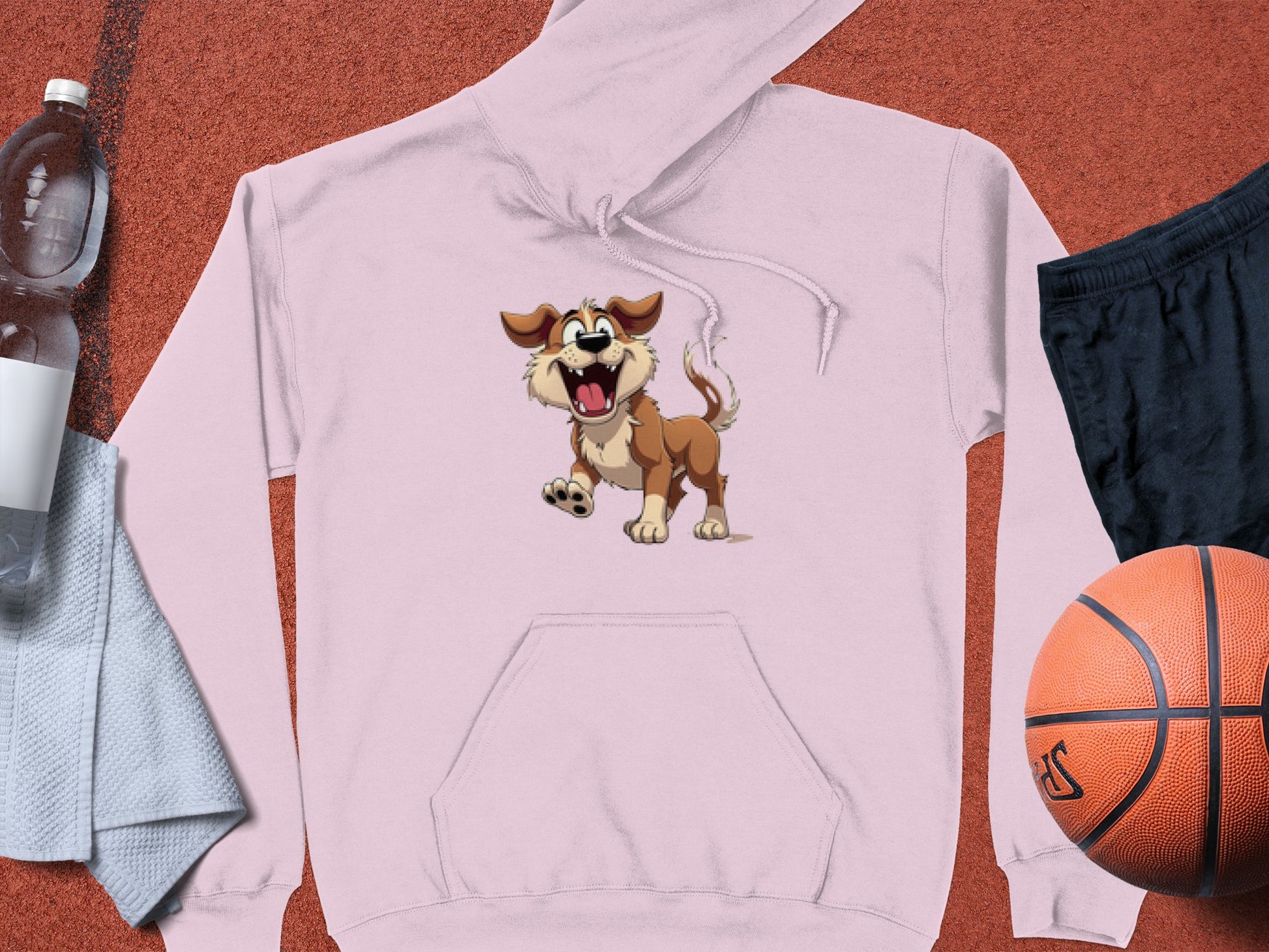 A light pink cartoon dog hoodie with a playful expression from Garment Graphics rests on a textured surface, accompanied by a water bottle, gray towel, black shorts, and an orange basketball—ideal for dog lovers who appreciate fun design hoodies and sporty activities.