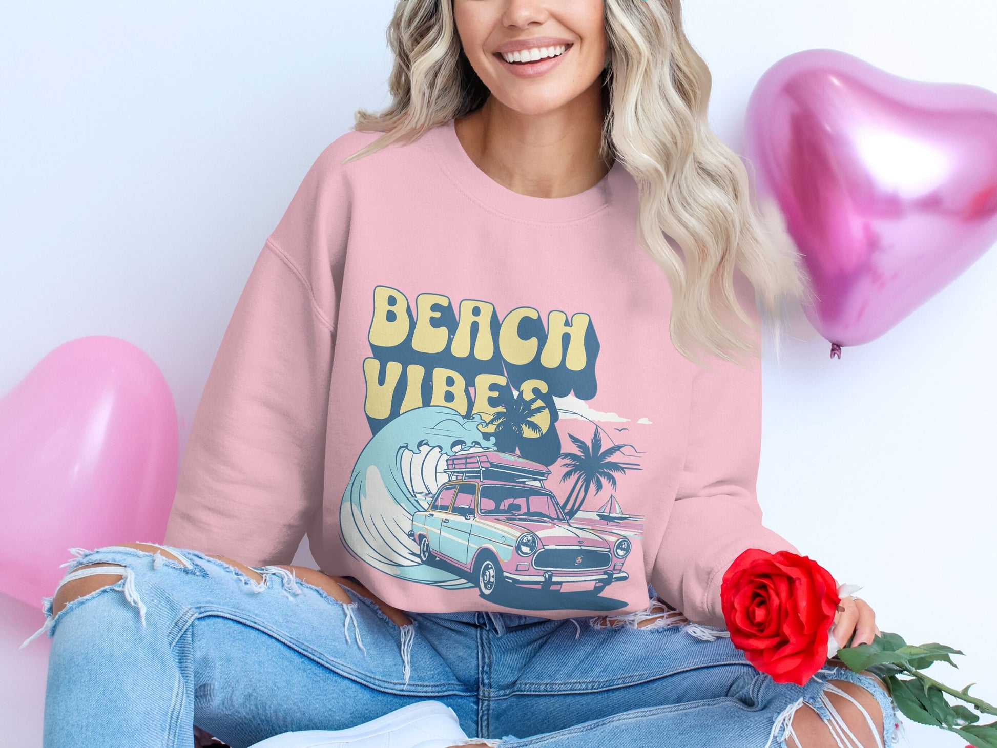 A person wears a Garment Graphics Beach Vibes pink sweatshirt with vintage car, palm trees, and wave graphics. They sit cross-legged in ripped jeans, holding a red rose while heart-shaped balloons float nearby, enhancing the retro beach vibe.