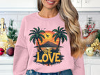 In a cozy holiday-decorated kitchen with white tiled walls, someone wears a Garment Graphics pink sweatshirt featuring two palm trees and a mountain against the One Love Sunset.