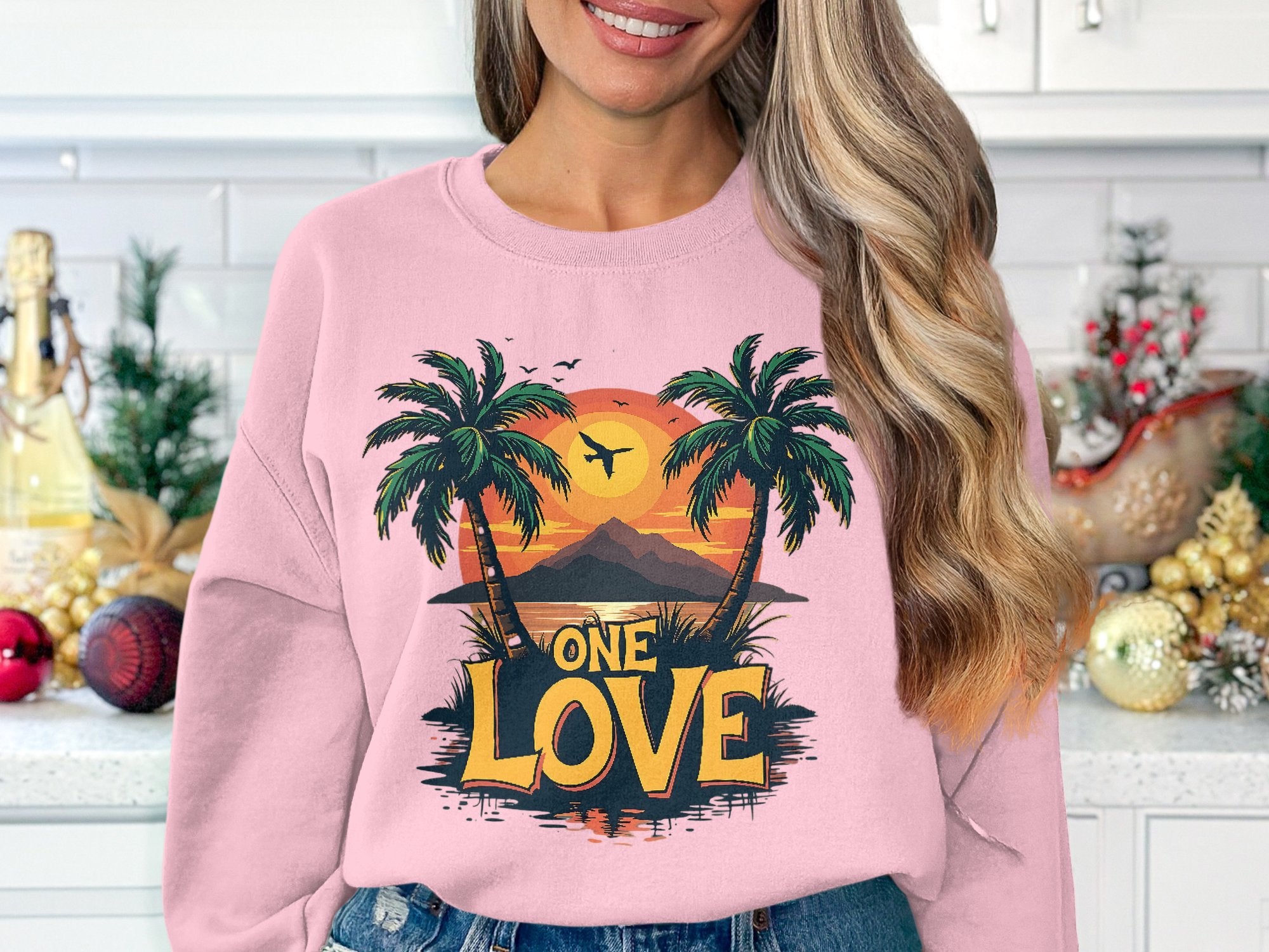 In a cozy holiday-decorated kitchen with white tiled walls, someone wears a Garment Graphics pink sweatshirt featuring two palm trees and a mountain against the One Love Sunset.