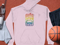 A retro light pink hoodie from Garment Graphics, featuring a colorful Woodstock 1969 graphic with a car and rainbow colors, is displayed on a textured surface beside a basketball, black shorts, water bottle, and light-colored towel.