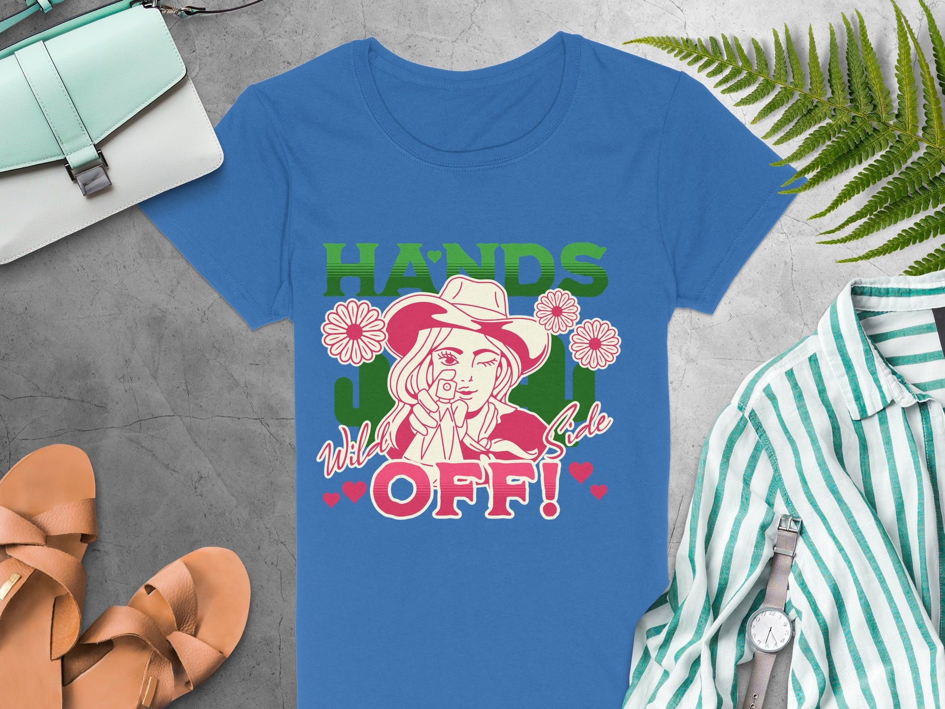 Garment Graphics offers a blue T-shirt with a bold design of a hat-wearing person holding a lasso, surrounded by flowers and Hands Off! Wild Side text. The cowboy graphic tee is styled alongside a handbag, sandals, striped shirt, and greenery on a gray backdrop.