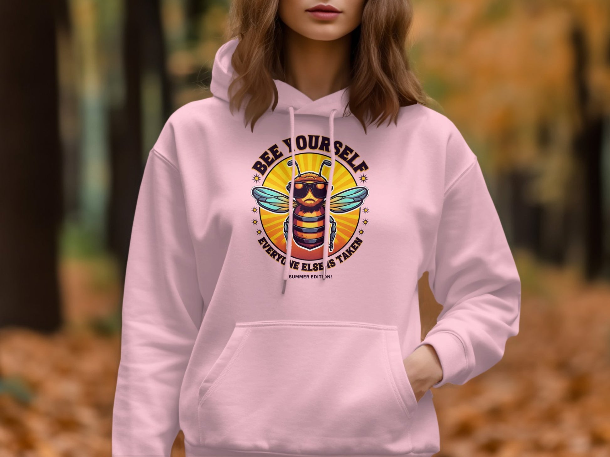 In a forest scene with autumn foliage, someone wears Garment Graphics vibrant pink Bee Yourself Hoodie, which showcases a bee graphic and the phrase Bee Yourself, Everyone Else is Taken, adding a playful flair to the natural setting.