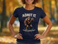 A person wears a navy Garment Graphics T-shirt showcasing a cartoon Bigfoot flashing a peace sign with the text Admit It Bigfoot, life would be boring without me, set against blurred autumn trees.