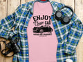 A pink Garment Graphics t-shirt with the Enjoy Vintage Ride design lies on a wooden surface, paired with a blue plaid shirt, headphones, a camera, and a white travel mug. Ideal for car enthusiasts, it features a classic car and reads Vintage Ride Community Since 1980, Aircooled.