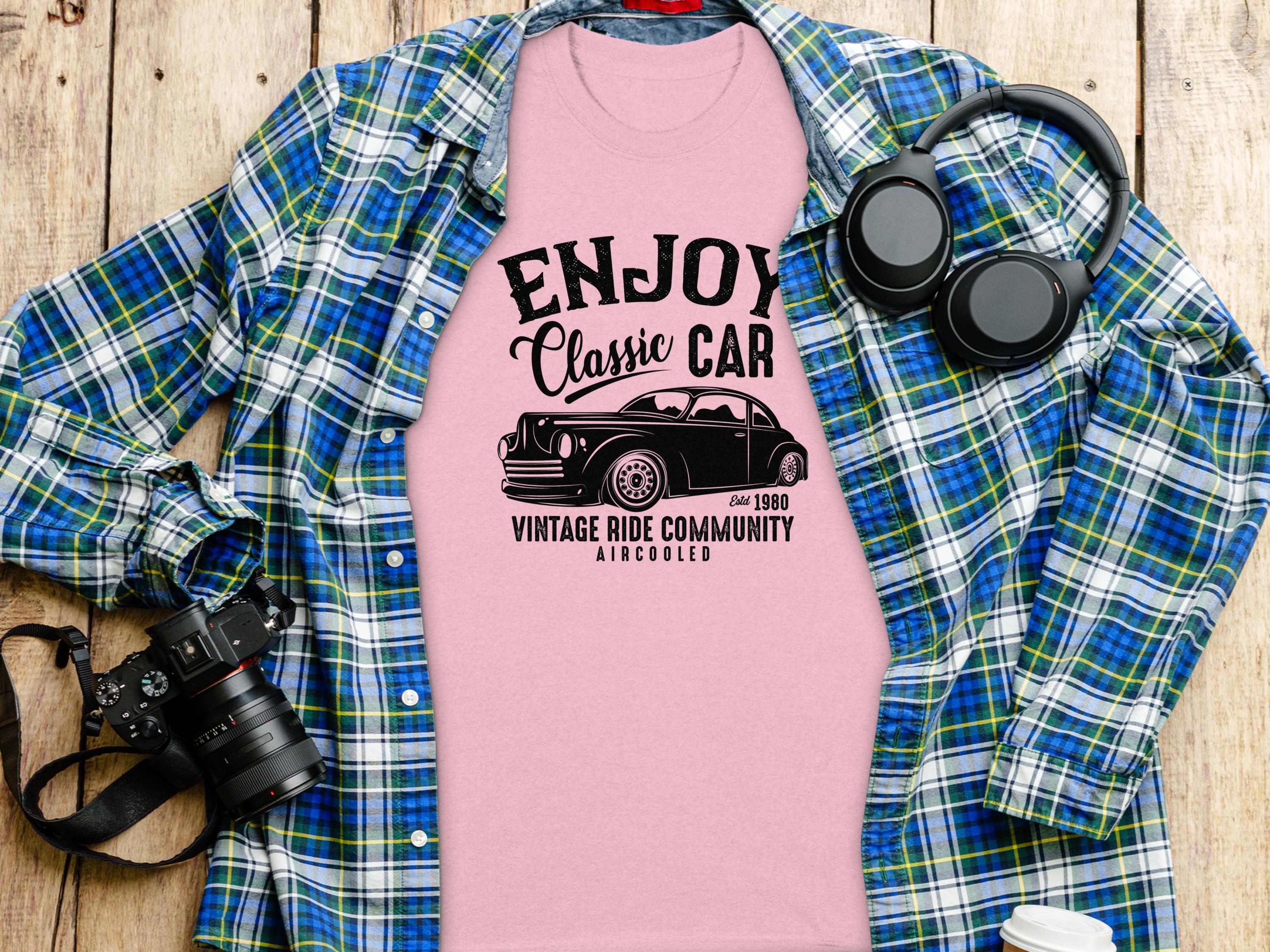 A pink Garment Graphics t-shirt with the Enjoy Vintage Ride design lies on a wooden surface, paired with a blue plaid shirt, headphones, a camera, and a white travel mug. Ideal for car enthusiasts, it features a classic car and reads Vintage Ride Community Since 1980, Aircooled.