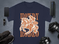 A Garment Graphics navy blue Rodeo T-shirt displays a cowgirl on horseback with stars and the text Howdy Rodeo and Not Beginners, ideal for enthusiasts, set among a smartphone, smartwatch, shoes, speaker, and dumbbells.