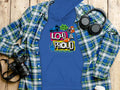 A Garment Graphics blue hoodie with Loud & Proud, colorful megaphone, and musical notes graphics lies under a blue plaid shirt on a wooden surface. Nearby are headphones, a camera, and a paper coffee cup. Its perfect for those ready to make some noise.