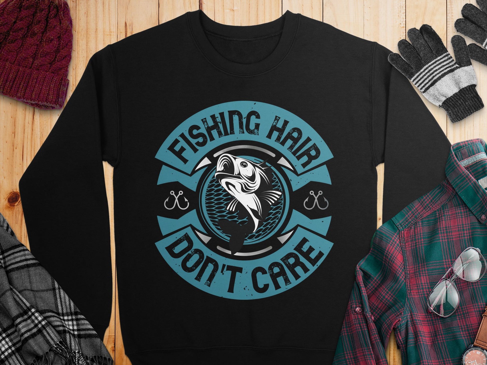 A black fishing sweatshirt by Garment Graphics features a circular fish graphic with Fishing Hair Dont Care in blue. Its styled with a maroon knit hat, plaid shirt, and black-and-white gloves on a wooden surface, perfect for fishing enthusiasts.