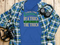 The Garment Graphics blue hoodie features a humorous quote: If an Englishman gets run over by a truck, he apologizes to the truck. Its paired with a blue and green plaid shirt, surrounded by headphones, a camera, coffee cup, and watch on a wooden surface.