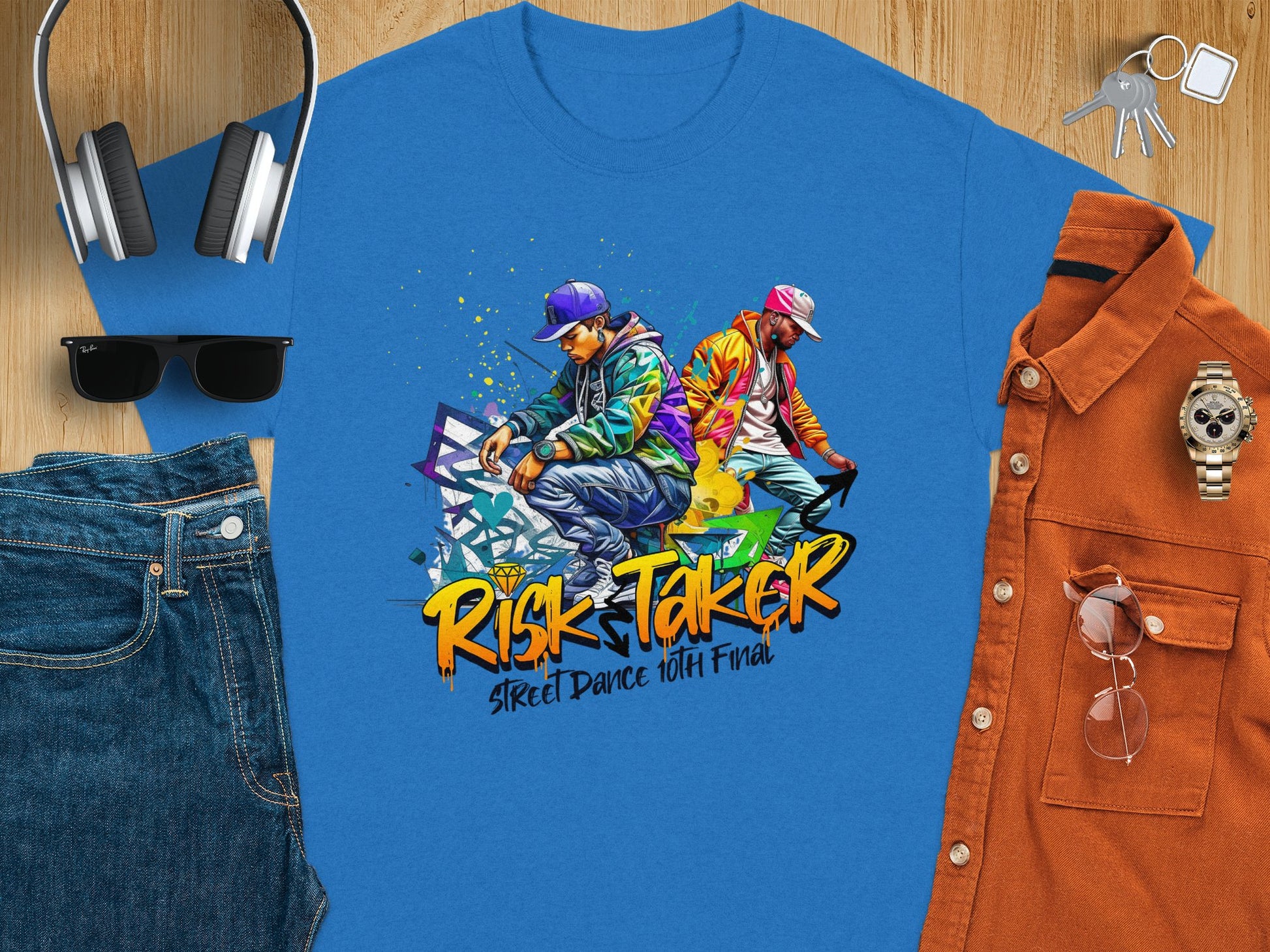 Garment Graphics blue t-shirt features a street dance culture graphic with Risk Taker 10th Final. Its styled with jeans, sunglasses, headphones, an orange button-up shirt, a wristwatch, and a smartphone on a wooden surface.