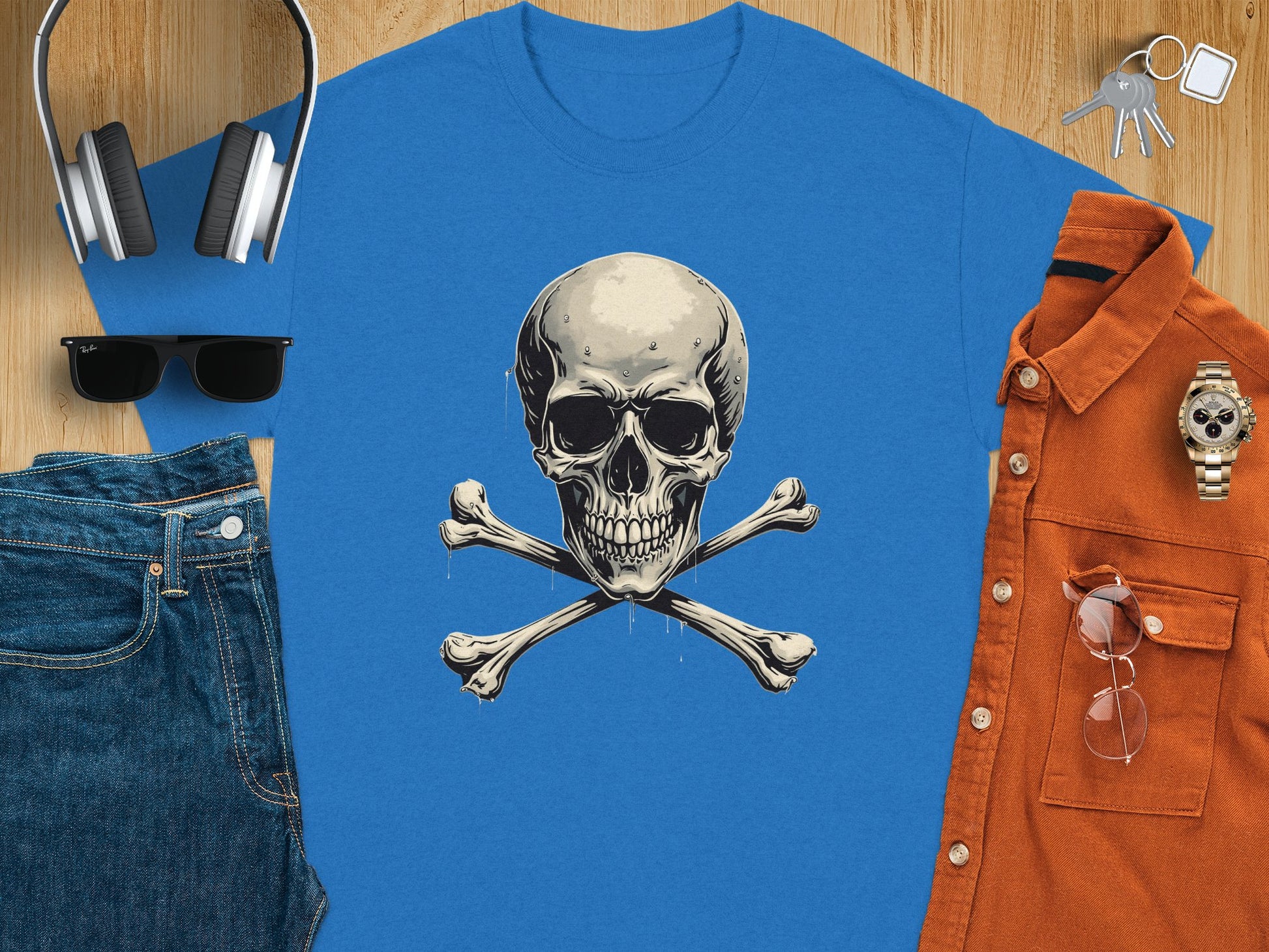 Garment Graphics blue T-shirt showcases a bold skull and crossbones design at its center—a standout fashion piece. Complementary essentials like jeans, sunglasses, headphones, a brown shirt, a wristwatch, and keys are scattered on the wood surface.