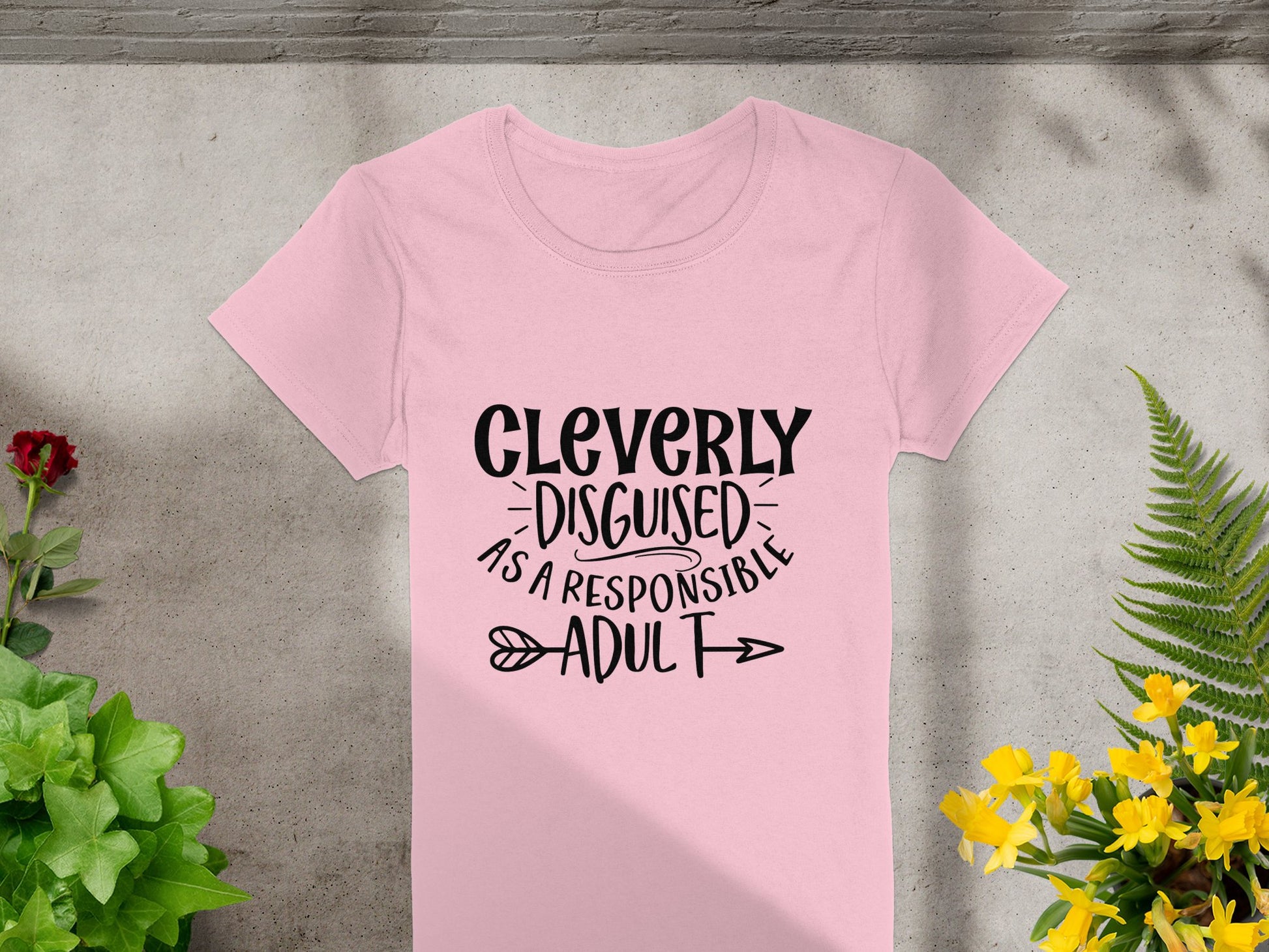 A pink semi-fitted T-shirt by Garment Graphics features Cleverly Disguised as a Responsible Adult in bold black with decorative arrows. Its beautifully displayed on a stone surface, surrounded by vibrant green plants and cheerful yellow flowers.