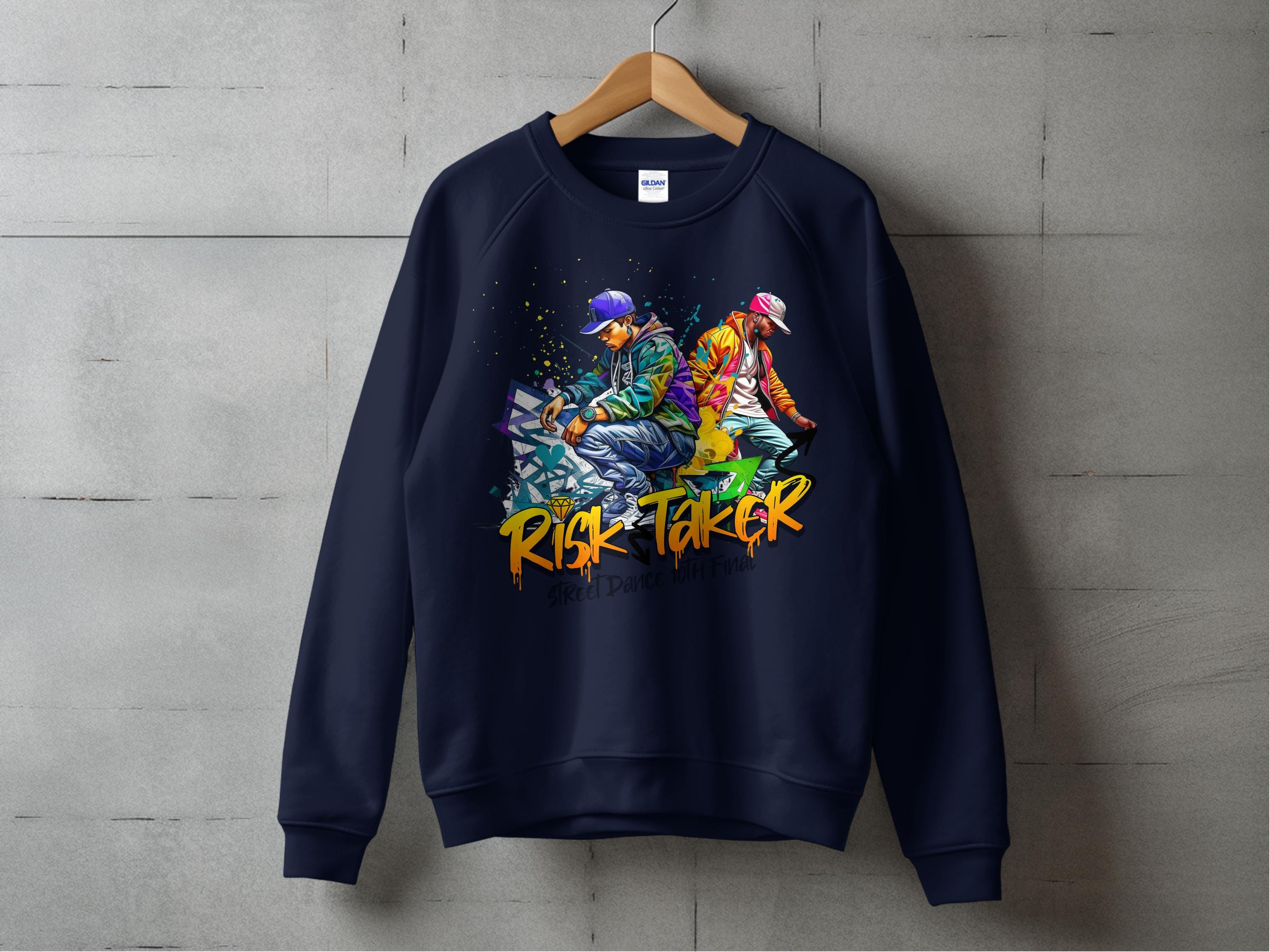 A navy sweatshirt by Garment Graphics hangs on a wooden hanger against a concrete wall, with street dance graphics of two people back-to-back. Risk Taker is boldly printed in orange below the image. The unisex design embodies urban style.